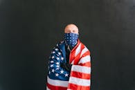 A man wrapped in a US flag wearing a mask stands against a dark backdrop, symbolizing resilience.